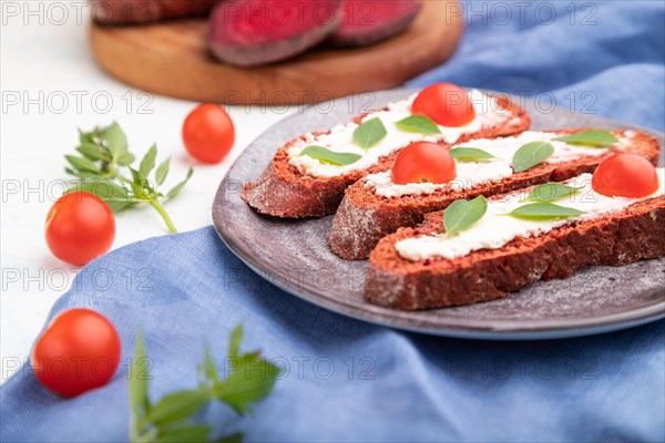Red beet bread sandwiches with cream cheese and tomatoes on white concrete background and blue linen textile. Side view, selective focus, close up