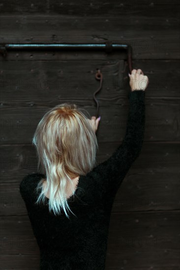 Back view of a blonde thin woman dressed in black reaching up to a rusty antique metal lock on a wooden door
