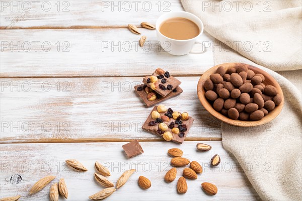 Almond in chocolate dragees in wooden plate and a cup of coffee on white wooden background and linen textile. Side view, copy space