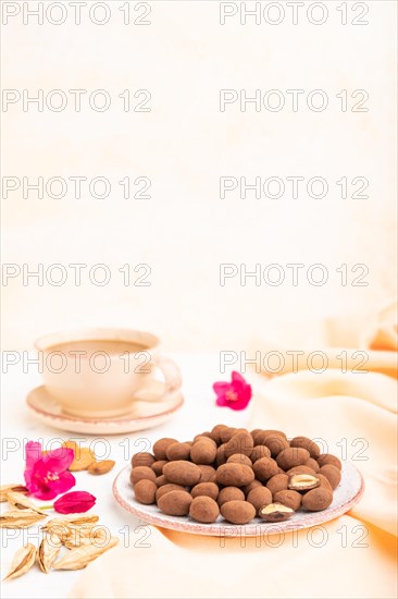 Almond in chocolate dragees on ceramic plate and a cup of coffee on white concrete background and orange linen textile. Side view, copy space, selective focus