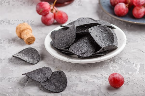 Black potato chips with charcoal, balsamic vinegar in glass, red grapes on a blue ceramic plate on a gray concrete background. side view, close up, selective focus