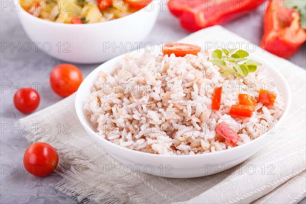 Unpolished rice porridge with stewed vegetables and oregano in white bowl on a gray concrete background and linen textile. side view, close up, selective focus