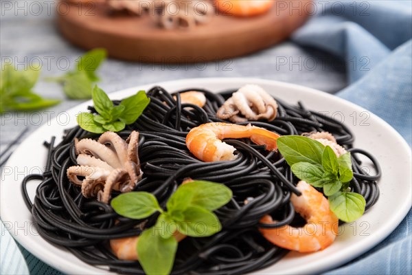 Black cuttlefish ink pasta with shrimps or prawns and small octopuses on gray wooden background and blue textile. Side view, close up, selective focus