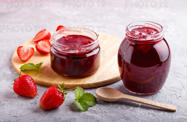 Strawberry jam in a glass jar with berries and leaves on gray concrete background. Homemade, close up, side view, selective focus