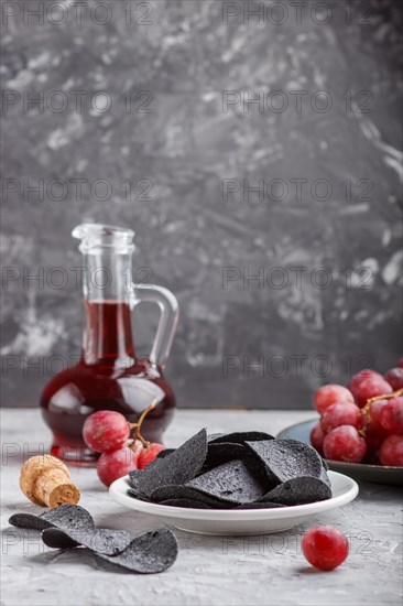 Black potato chips with charcoal, balsamic vinegar in glass, red grapes on a blue ceramic plate on a gray concrete background. side view, close up, selective focus, copy space