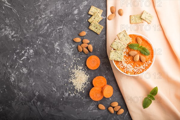 Carrot cream soup with sesame seeds, almonds and snacks in white bowl on a black concrete background with orange textile. top view, flat lay, copy space
