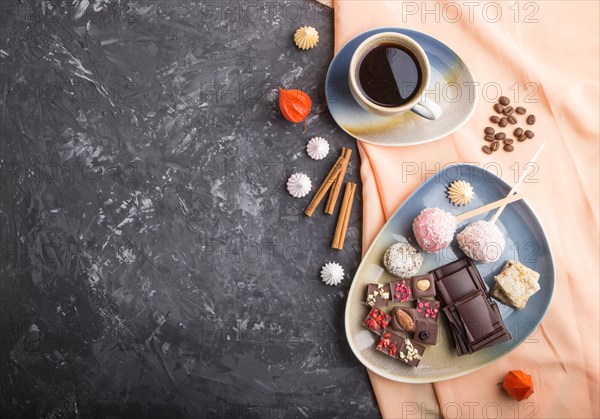 A pieces of homemade chocolate with coconut candies and a cup of coffee on a black concrete background and orange textile. top view, flat lay, copy space