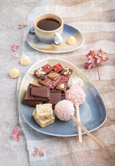 A pieces of homemade chocolate with coconut candies and a cup of coffee on a gray concrete background and blue and brown textile. side view, close up