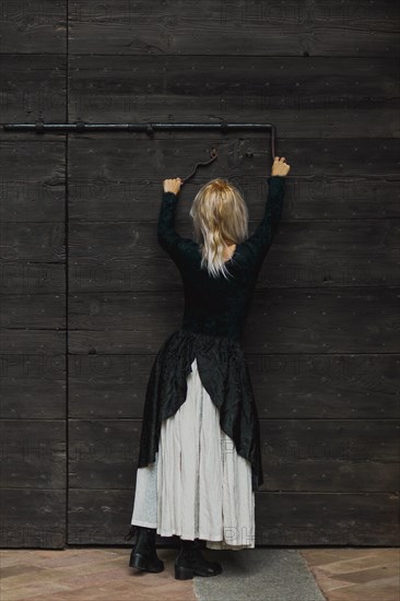 Full body shot of a young blonde caucasian woman reaching an antique lock of a wooden door above her head, wearing a black top and white skirt and a furry shirt