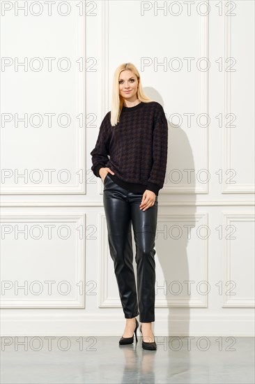 A stylish woman standing against a white, panelled wall. She is dressed in a dark, patterned sweater and sleek black leather pants, complemented by classic black heels