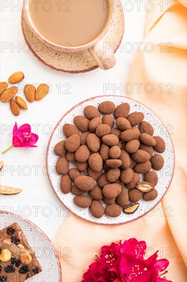 Almond in chocolate dragees on ceramic plate and a cup of coffee on white concrete background and orange linen textile. Top view, close up, flat lay