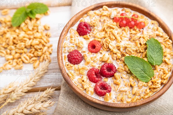 Wheat flakes porridge with milk, raspberry and currant in wooden bowl on white wooden background and linen textile. Side view, close up, selective focus