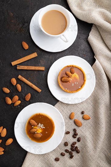 Sweet tartlets with almonds and caramel cream with cup of coffee on a black concrete background and linen textile. top view, flat lay, close up