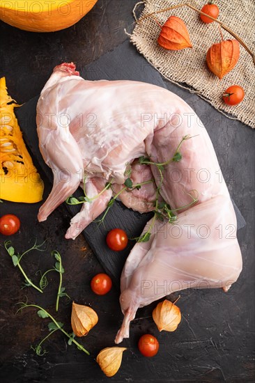 Whole raw rabbit with pumpkin, tomatoes, pea sprouts on a black concrete background and linen textile. Top view, flat lay, close up