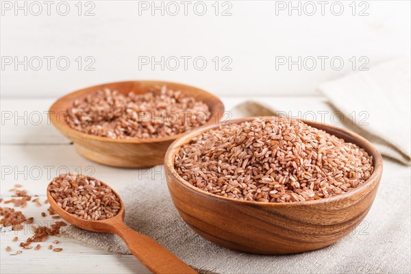 Two wooden bowls with unpolished brown rice and wooden spoon on a white wooden background and linen textile. Side view, close up, selective focus