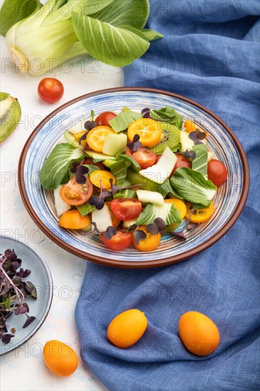 Vegetarian salad of pac choi cabbage, kiwi, tomatoes, kumquat, microgreen sprouts on a white concrete background and blue linen textile. Side view, close up