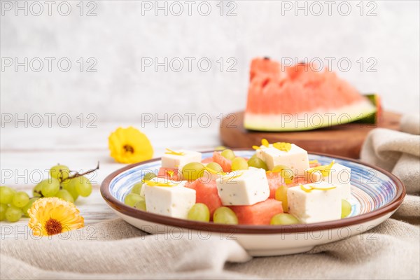 Vegetarian salad with watermelon, feta cheese, and grapes on blue ceramic plate on white wooden background and linen textile. Side view, close up, selective focus