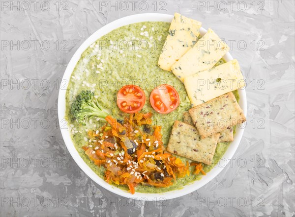 Green broccoli cream soup with crackers and cheese in white bowl on a gray concrete background. top view, flat lay, close up