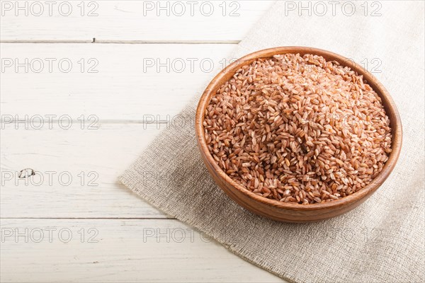 Wooden bowl with unpolished brown rice on a white wooden background and linen textile. Side view, copy space