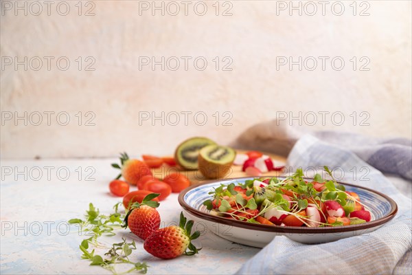 Vegetarian fruits and vegetables salad of strawberry, kiwi, tomatoes, microgreen sprouts on white concrete background and linen textile. Side view, copy space, selective focus
