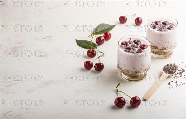 Yoghurt with cherries, chia seeds and granola in glass with wooden spoon on white wooden background. side view, copy space