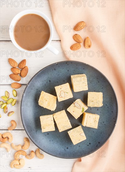 Traditional indian candy soan papdi in a blue ceramic plate with almond, pistache, cashew and a cup of coffee on a white wooden background with orange textile. top view, flat lay, close up