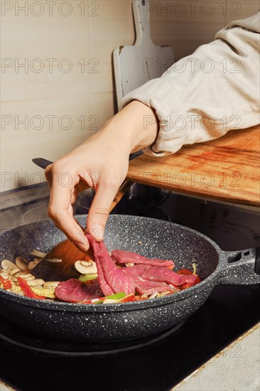 Unrecognizable woman adds thin slices of beef to pan with vegetable dressing