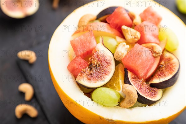 Vegetarian fruit salad of watermelon, grapes, figs, pear, orange, cashew on slate board on a black concrete background. Side view, close up, selective focus