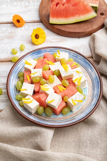 Vegetarian salad with watermelon, feta cheese, and grapes on blue ceramic plate on white wooden background and linen textile. Side view, close up