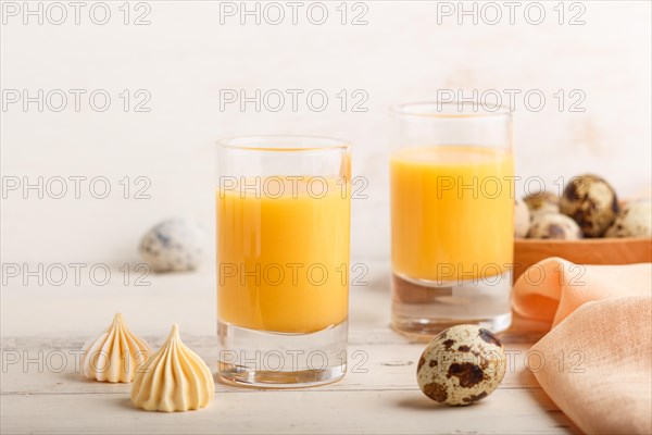 Sweet egg liqueur in glass with quail eggs and meringues on white wooden background. Side view, close up, high key