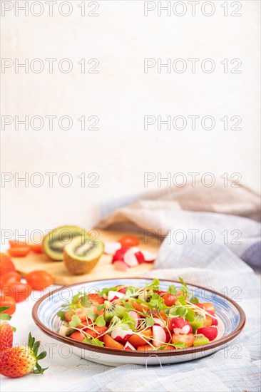 Vegetarian fruits and vegetables salad of strawberry, kiwi, tomatoes, microgreen sprouts on white concrete background and linen textile. Side view, copy space, selective focus