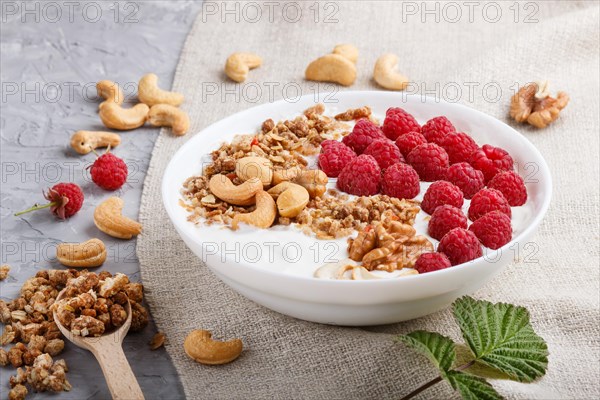 Yoghurt with raspberry, granola, cashew and walnut in white plate with wooden spoon on gray concrete background and linen textile. side view, close up