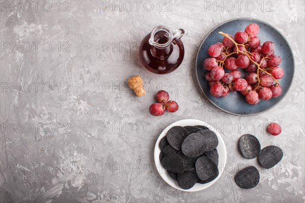 Black potato chips with charcoal, balsamic vinegar in glass, red grapes on a blue ceramic plate on a gray concrete background. Top view, flat lay, copy space
