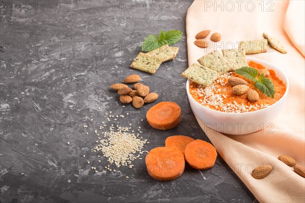 Carrot cream soup with sesame seeds, almonds and snacks in white bowl on a black concrete background with orange textile. side view, copy space