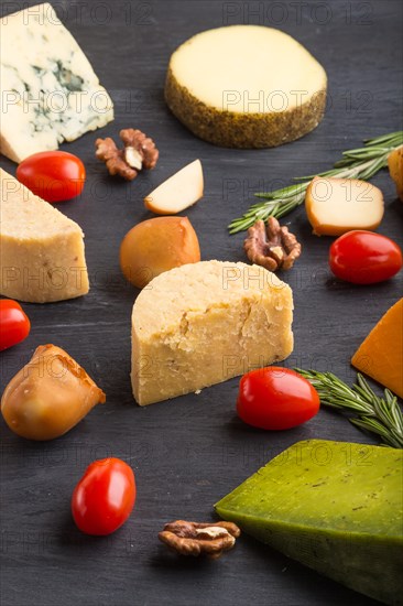 Set of different types of cheese with rosemary and tomatoes on a black wooden background. Side view, close up