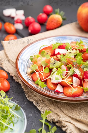 Vegetarian fruits and vegetables salad of strawberry, kiwi, tomatoes, microgreen sprouts on black concrete background and linen textile. Side view, close up, selective focus