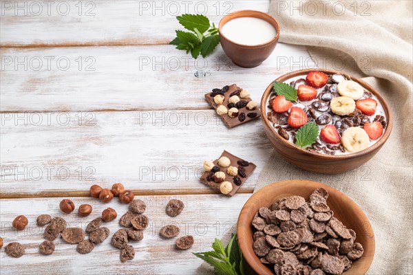 Chocolate cornflakes with milk and strawberry in wooden bowl on white wooden background and linen textile. Side view, copy space