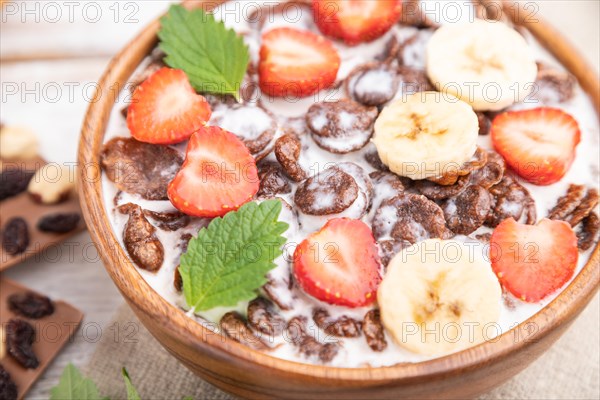 Chocolate cornflakes with milk and strawberry in wooden bowl on white wooden background and linen textile. Side view, close up, selective focus