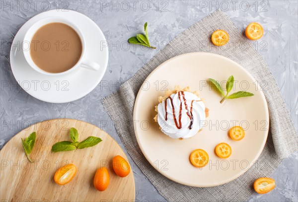 Cake with whipped egg cream on a light brown plate with kumquat slices and mint leaves on a gray concrete background with linen napkin and cup of coffee. copy space, flat lay, top view