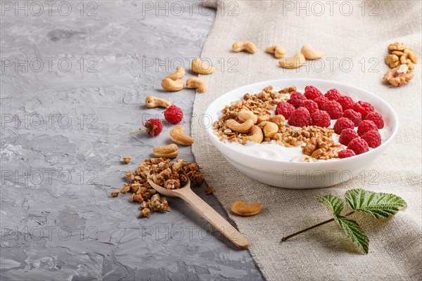 Yoghurt with raspberry, granola, cashew and walnut in white plate with wooden spoon on gray concrete background and linen textile. side view, copy space