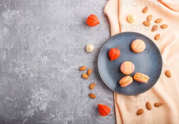 Orange macarons or macaroons cakes on blue ceramic plate on a gray concrete background and orange textile. Flat lay, top view, copy space