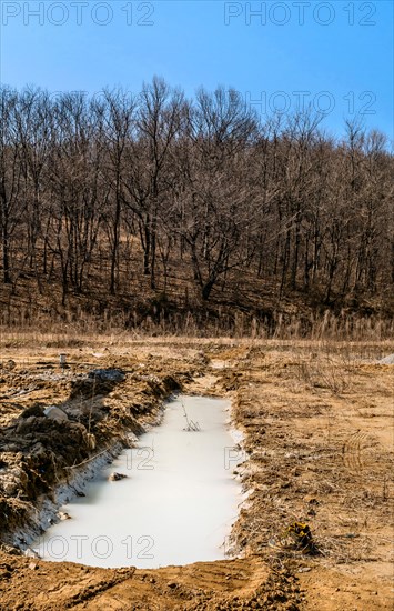 Hiking shoes left beside man made ditch filled with milky water in wilderness with leafless trees in background in South Korea