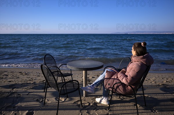 Young woman looking at the sea, lonely, alone, coat, table and chairs, beach bar, beach, Peraia, also Perea, Thessaloniki, Macedonia, Greece, Europe