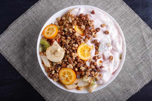 Vegetarian salad of bananas, apples, pears, kumquats, kiwi with granola and yogurt on black woodem background, top view