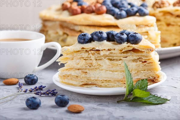 Homemade layered Napoleon cake with milk cream. Decorated with blueberry, almonds, walnuts, hazelnuts, mint on a gray concrete background and cup of coffee. side view, close up, selective focus