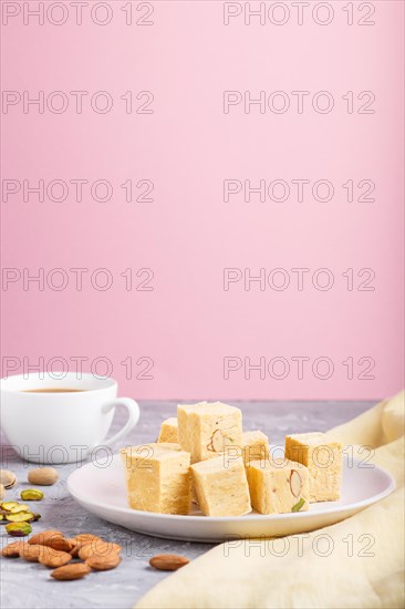 Traditional indian candy soan papdi in white plate with almond, pistache and a cup of coffee on a gray and pink background with yellow textile. side view, close up, selective focus