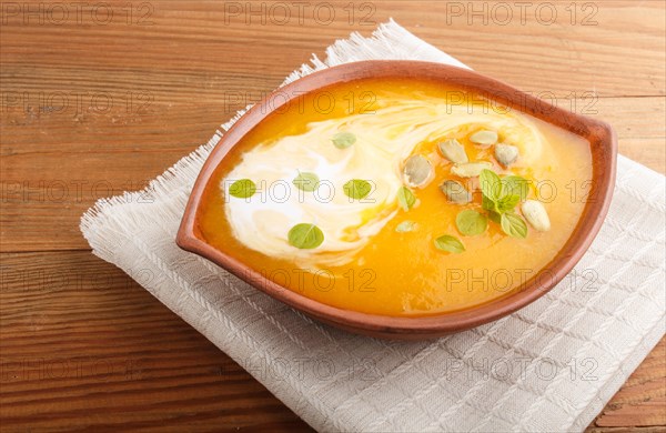 Traditional pumpkin cream soup with seeds in clay bowl on a brown wooden background with linen napkin. side view, close up