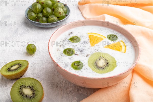 Yogurt with kiwi, gooseberry, chia in ceramic bowl on gray concrete background and orange linen textile. Side view, close up