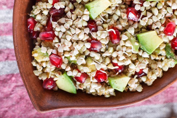 Salad of germinated buckwheat, avocado, walnut and pomegranate seeds in clay plate on white wooden background. Top view, close up, selective focus