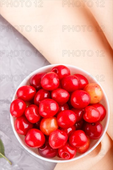 Fresh red sweet cherry in white bowl on gray background with orange textile. top view, flat lay